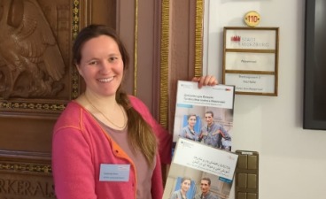 KAUSA-Mitarbeiterin präsentiert mehrsprachige Elternratgeber Auf dem Bild ist eine lächelnde Frau zu sehen, die Broschüren präsentiert.