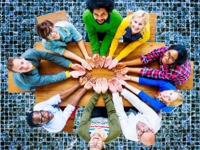 Mehrere Menschen unterschiedlicher ethnischer Abstammung sitzen an einem runden Tisch. Ihre Hände treffen sich in der Mitte.