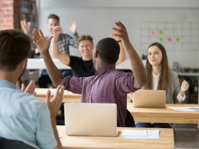 Erwachsene Schüler*innen sitzen im Unterrichtsraum. Es ist eine gute Stimmung zu erkennen. Schüler reißen die Arme hoch oder klatschen.