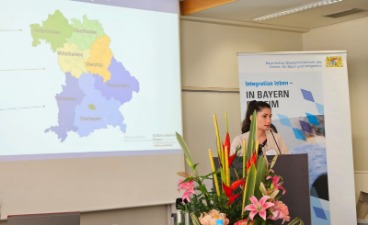 Eine Projektmitarbeiterin aus München steht hinter einem Pult. Neben ihr ist eine Leinwand mit einer Landkarte Bayerns zu sehen.