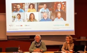 Zwei Frauen sitzen vor einer Leinwand mit dem Titelbild der Kausa-Landesstelle Bayern