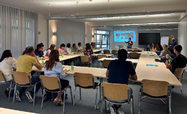 Informationsveranstaltung für Akademiker*innen Gäste der Informationsveranstaltung sitzen in einem großen Raum an Tischen, die in U-Form angeordnet sind. Sie sind überwiegend von hinten zu sehen und schauen zu einer Präsentation nach vorne.
