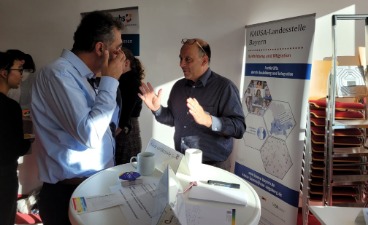 Infostand beim Netzwerktreffen zum "Onboarding" Ein Mann spricht an einem Infostand mit einem anderen Mann. Im Hintergrund ist das Plakat der KAUSA-Landesstelle zu sehen.