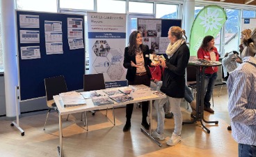 Infostand auf der Netzwerkmesse Oberallgäu Eine Frau spricht an einem Infostand mit einer anderen Frau. Im Hintergrund ist das Plakat der KAUSA-Landesstelle zu sehen.